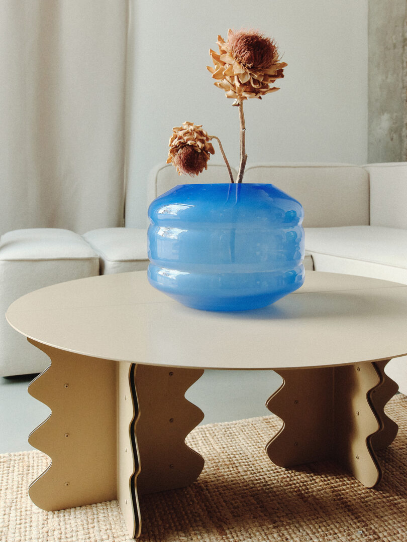 Round table with a large blue vase holding dried flowers. Beige sofa and curtain in the background.