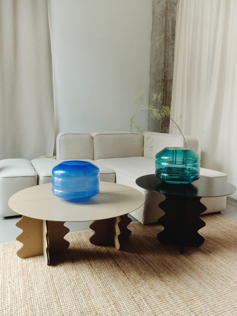 Two modern coffee tables with wavy legs, one white with a blue glass vase and one black with a teal glass vase, in front of a beige sectional sofa on a woven rug.