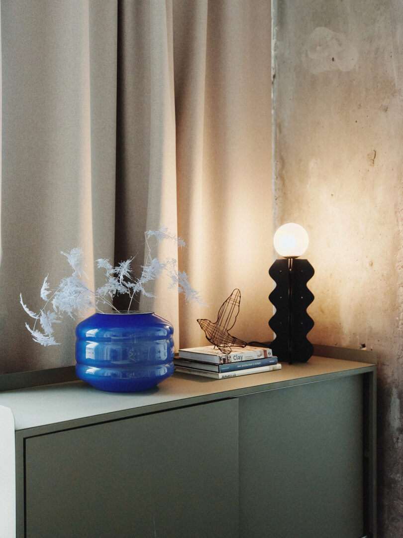 A minimalist scene with a blue vase, a metal butterfly sculpture, books, and a wavy black lamp with a round bulb on a cabinet beside beige curtains.