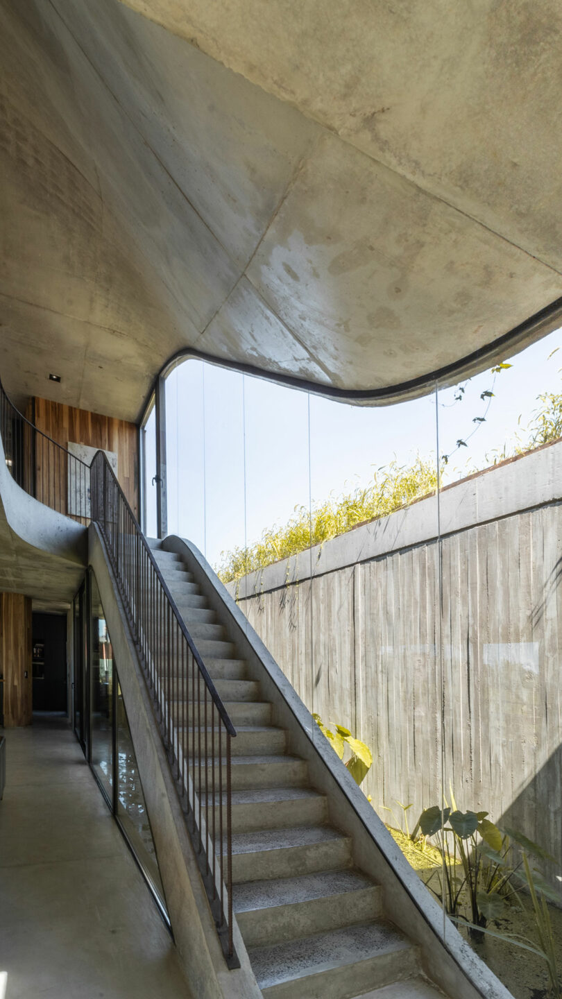 Concrete staircase with a sleek metal railing leading to an upper floor. Large glass walls offer an outside view with plants visible along the top edge of the wall.