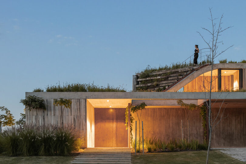 A modern concrete building with greenery on top. A person stands on the rooftop stairs. The front is illuminated, and it's surrounded by plants. Sky is clear with some clouds.