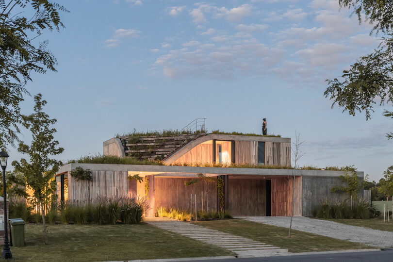 A modern two-story house with a green roof and wooden accents, set in a landscaped yard with a path leading to the entrance. A person stands on the roof.