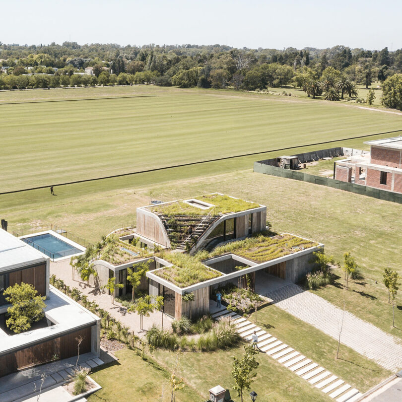 Modern house with green roofs and a pool, surrounded by grass and trees, next to a large open field.