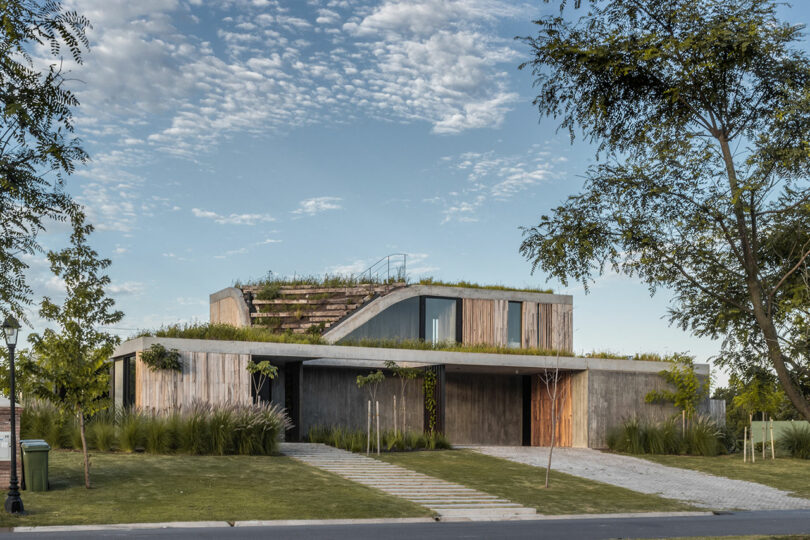 Modern house with a green roof and wooden exterior, surrounded by grass and trees under a cloudy sky.