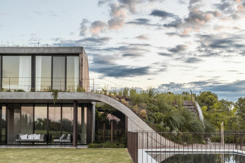 Modern house with large glass windows, green roof, and nearby grassy area, set against a cloudy sky backdrop.