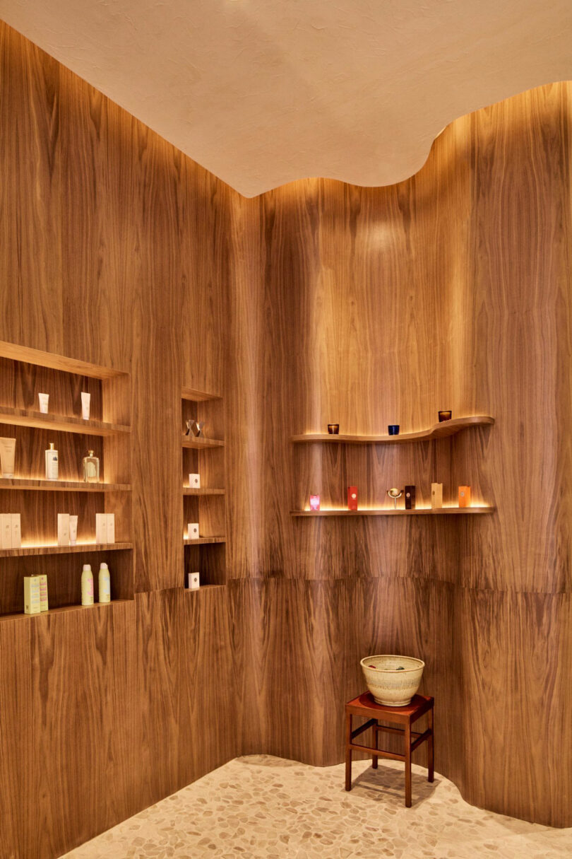 A minimalist interior with wooden walls and shelves holding various products. A small stool with a bowl is placed against the wall. Stone-patterned flooring complements the setting.