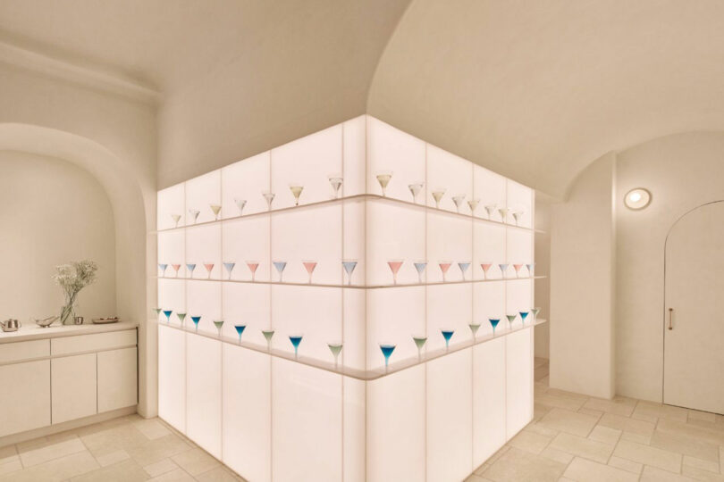 Minimalist room with a white display case filled with evenly spaced pastel-colored vases. Soft lighting highlights the geometric design. A vase with flowers is on a side cabinet.