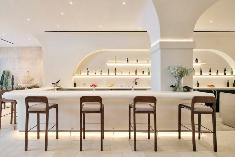 A minimalist bar with a white counter, wooden stools, and bottles displayed on backlit shelves against a light-colored wall.