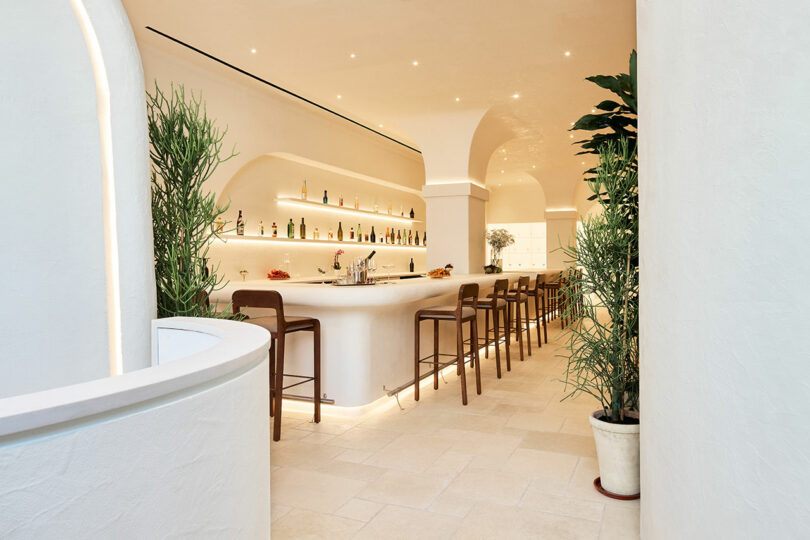 Sleek modern bar with high stools, illuminated shelves with bottles, arched alcoves, and potted plants in a minimalist white setting.