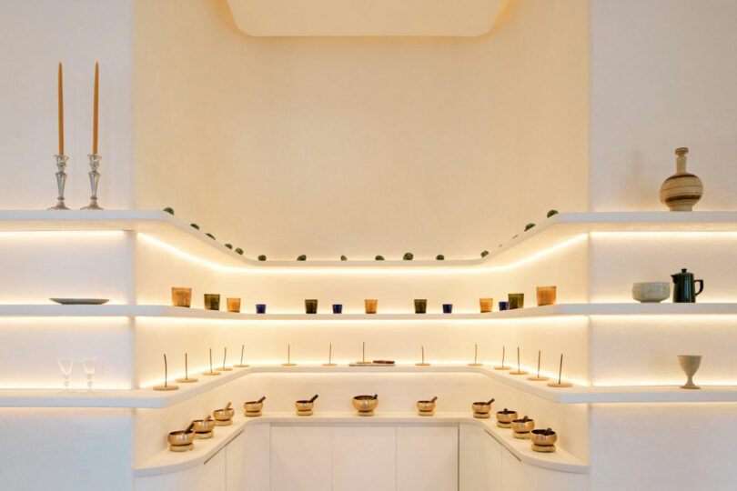 A minimalist, well-lit display of various cups, bowls, and decorative items on tiered white shelves.