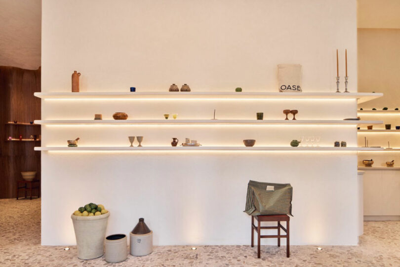 Minimalist interior with white walls and illuminated shelves displaying small decorative items. A chair with a cloth is placed against the wall, and pots are on the floor.