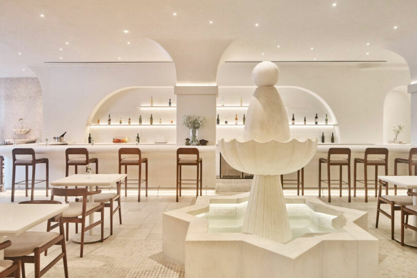 Elegant modern interior with a central white stone fountain, surrounded by tables and chairs. A sleek bar with shelves of bottles lines the back wall under minimal archways.