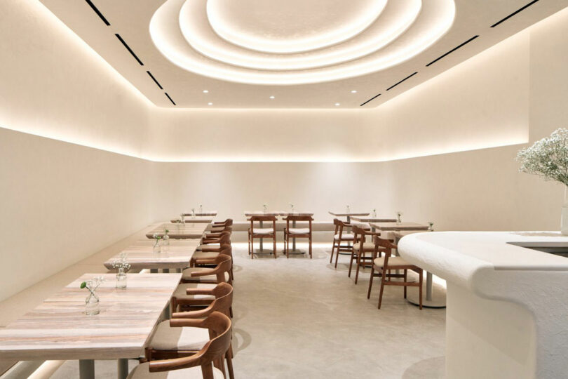 Minimalist cafe interior with wooden tables and chairs, beige walls, and ceiling lights. Tables have small vases with flowers.