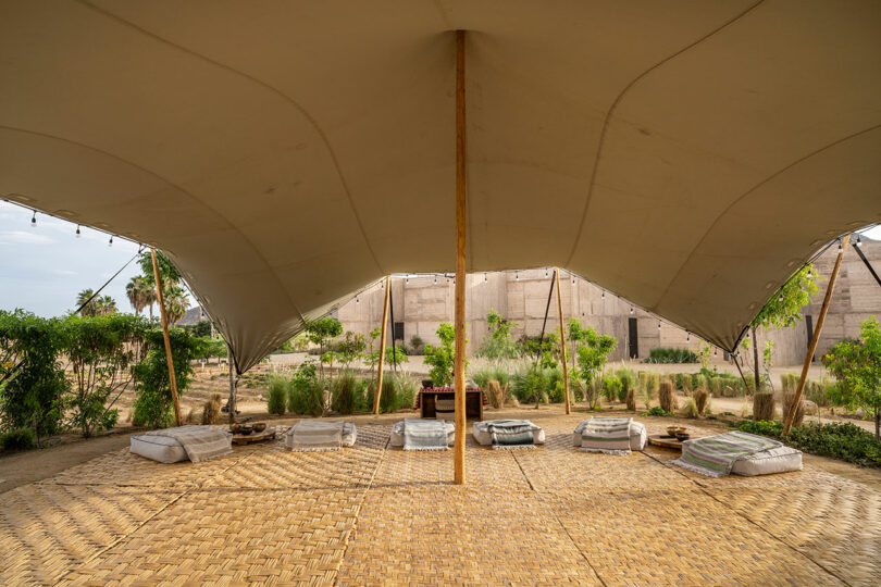 An open-sided tent structure with a woven mat floor, cushions arranged in a circle, and surrounding greenery in an outdoor setting.