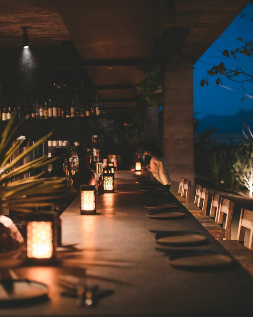 Long dining table with place settings and lit lanterns, set in a dimly lit restaurant. Shelves with bottles are on the side, and plants decorate the space.