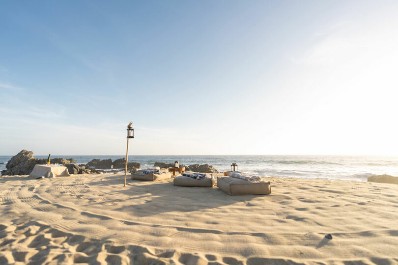 Beach setting with sandy ground, cushions, and lanterns near the ocean. Rocks are visible in the background under a clear sky.