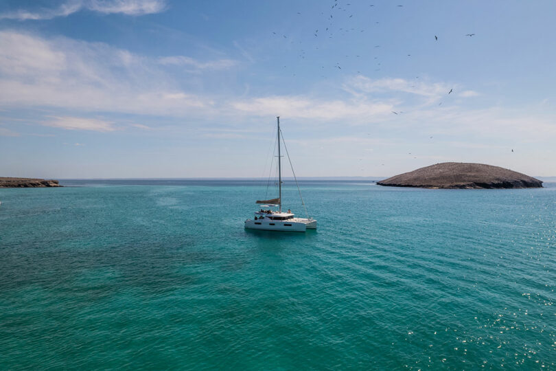 A sailboat floats on calm, turquoise waters with a rocky island in the background and birds flying overhead.