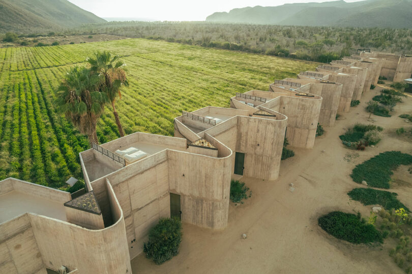 Aerial view of a row of identical, geometric concrete buildings beside a lush green field with mountains in the background.