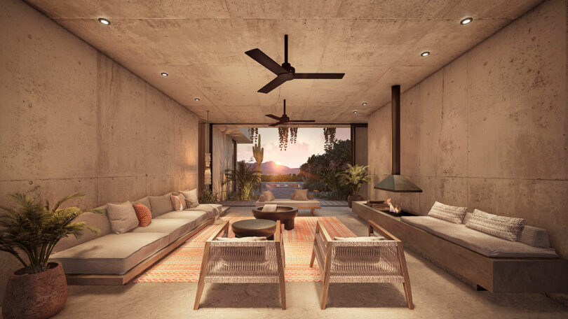 Minimalist living room with concrete walls, ceiling fans, sofas, and a tropical view of sunset through a large open window. Modern decor with plants and a central rug.