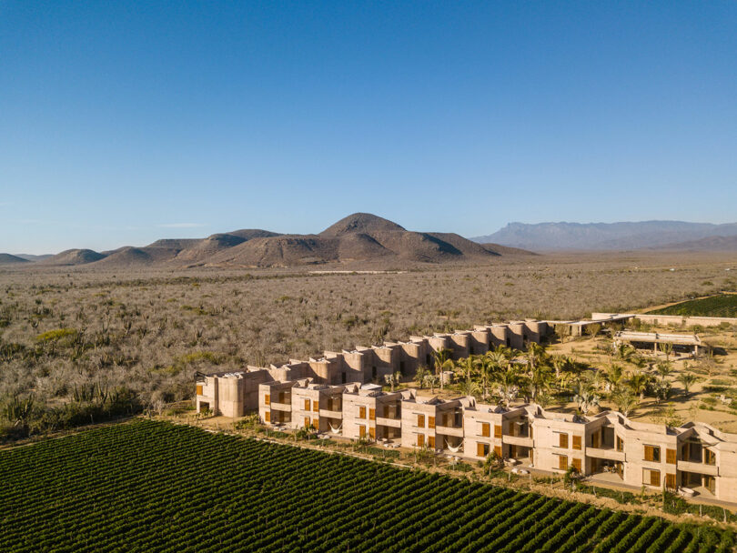 Aerial view of a series of beige buildings with palm trees in front, located on the edge of a field, with flat-topped mountains and open desert landscape in the background under a clear blue sky.