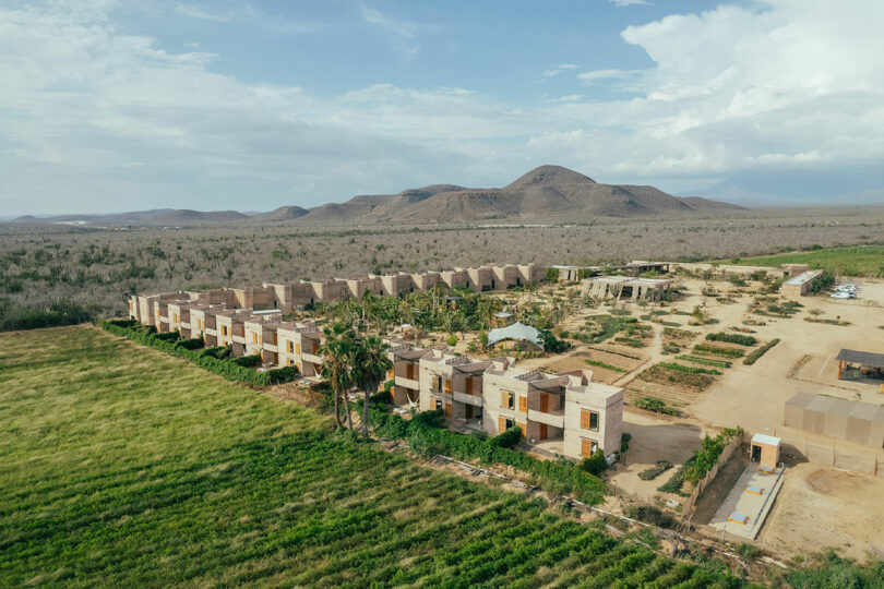 Aerial view of a rural landscape with a series of modern, rectangular buildings, adjacent fields, and distant mountains under a partly cloudy sky.