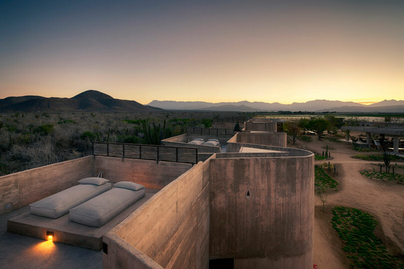 Rooftop view of an adobe-style building with two beds, surrounded by desert landscape and mountains at sunset.