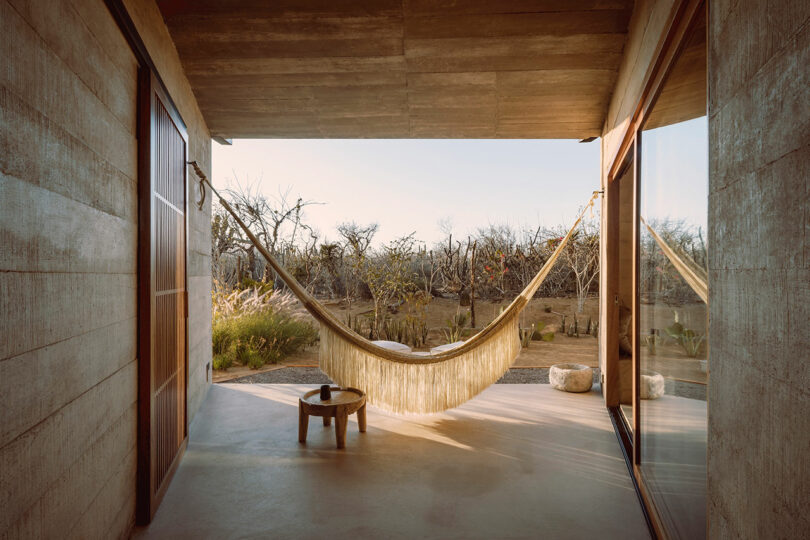 A covered patio with a hammock and a small round table, overlooking a dry landscape with sparse trees and shrubs.