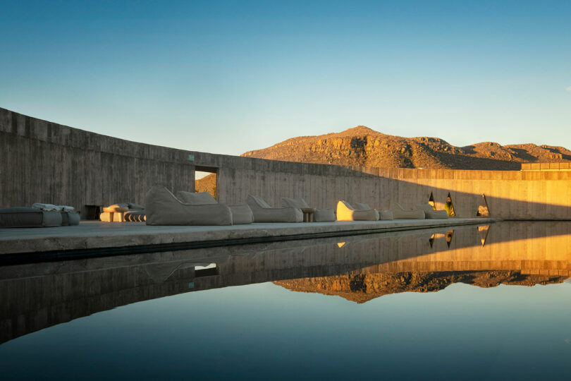 Modern outdoor seating area with beige couches along a wall next to a reflective water surface. Mountains are visible in the background under a clear blue sky.