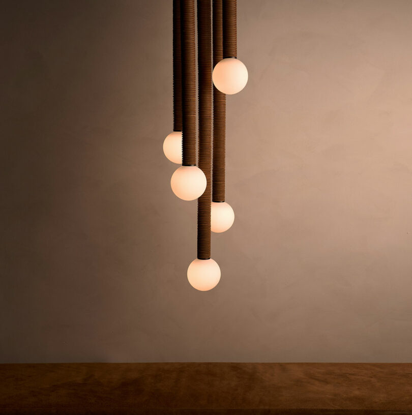 The Pell chandelier by Studio Luddite features a stunning design of six glowing round bulbs suspended from textured cables, creating an elegant contrast against the plain wall.