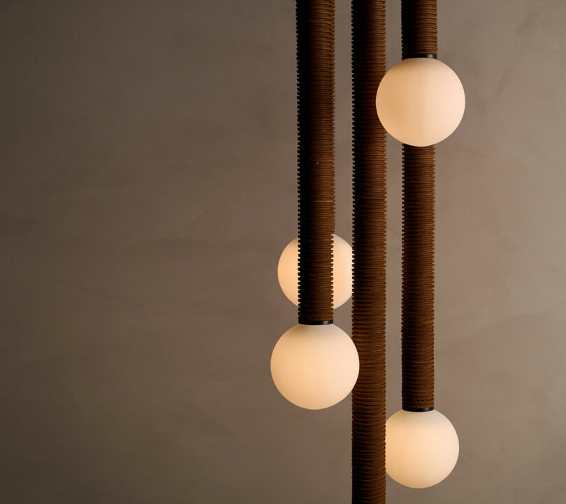 The Pell Chandelier by Studio Luddite showcases five illuminated spherical bulbs gracefully suspended from textured, vertical rods against a plain background.