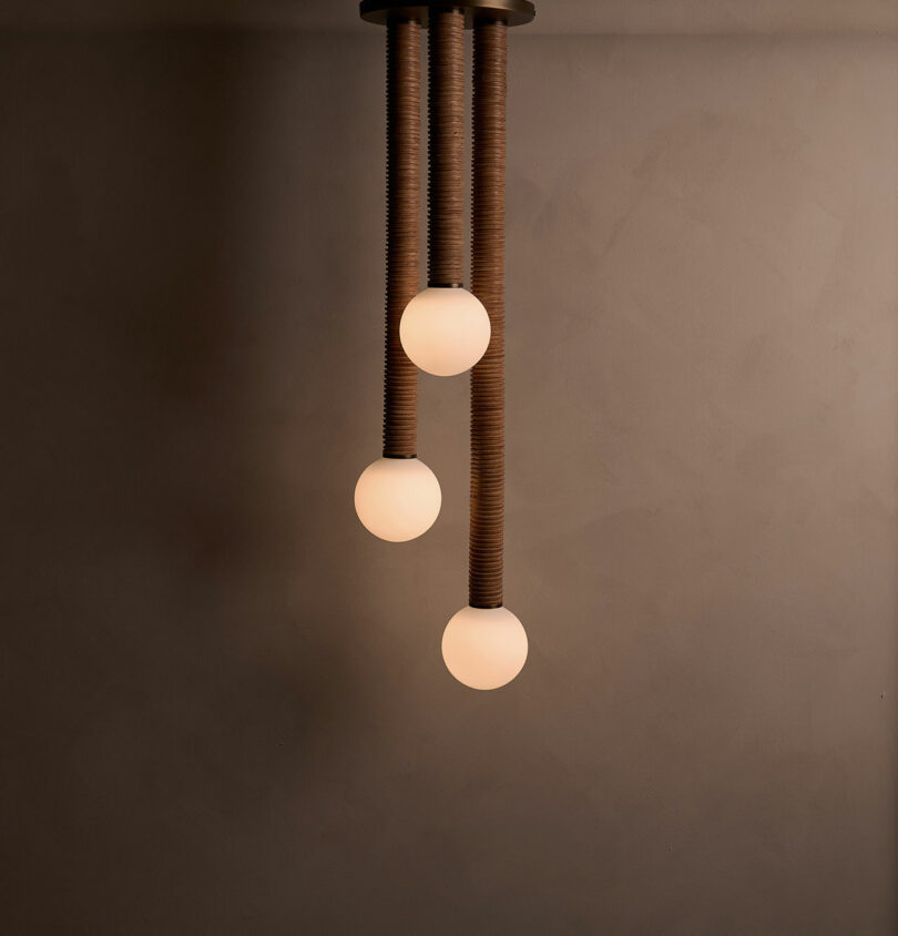 The Pell Chandelier from Studio Luddite is a modern pendant light fixture featuring three spherical bulbs elegantly suspended from textured cords against a plain wall.