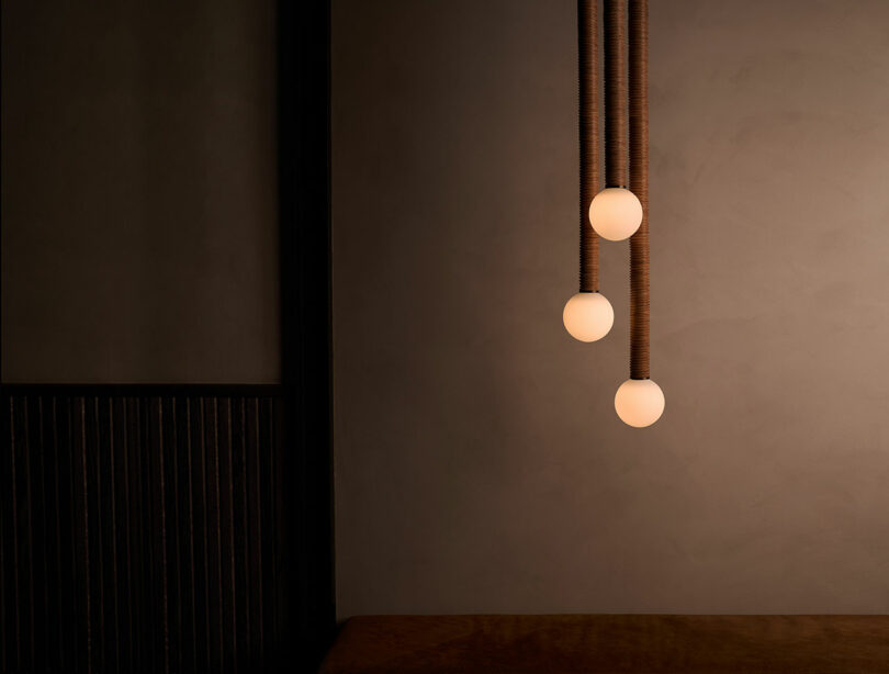 Three spherical pendant lights from Studio Luddite hang at varying heights against a minimalist dark wall.