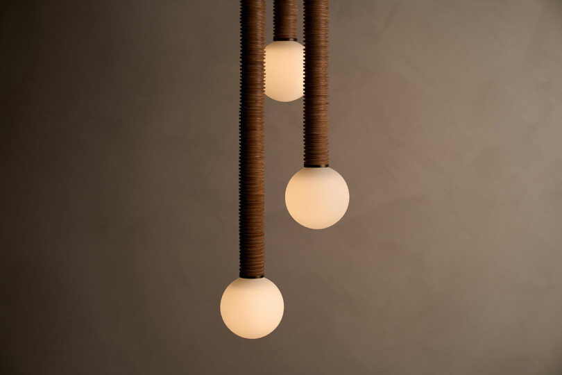 Three pendant lights with round, frosted bulbs hang from brown, textured cords against a gray background, embodying the exquisite craftsmanship of Studio Luddite.