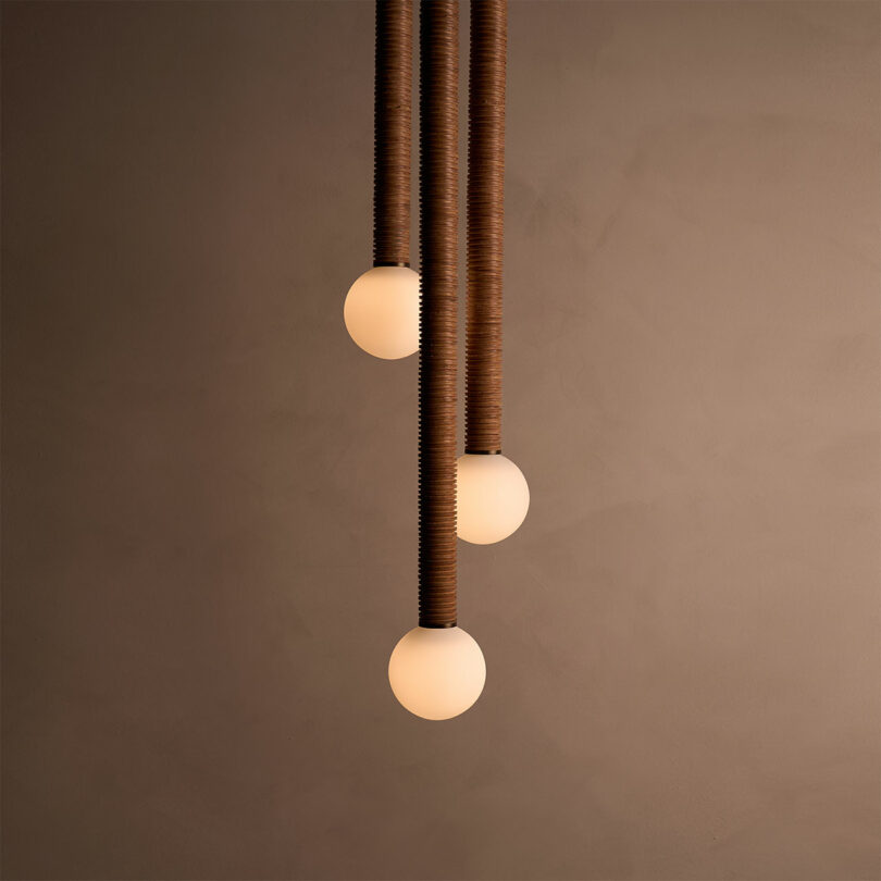 A trio of spherical pendant lights with wooden textured cables, artfully crafted by Pell Chandelier, hang elegantly against a plain brown wall.