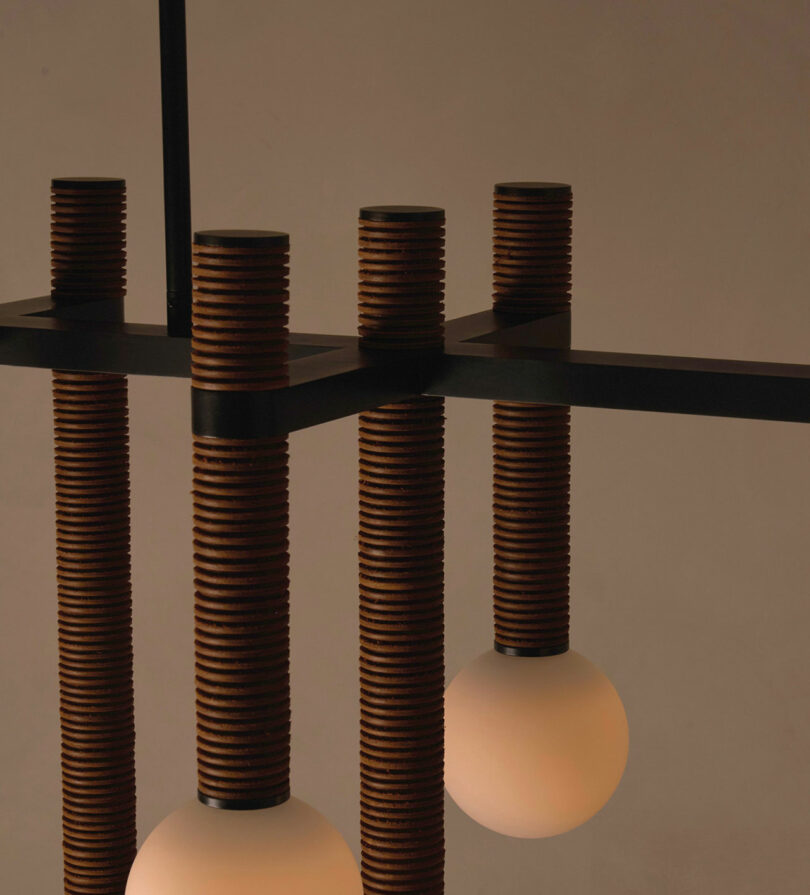 A modern light fixture by Studio Luddite features cylindrical wooden supports and spherical light bulbs, set against a neutral background. This Pell Chandelier adds a touch of contemporary elegance to any space with its unique design and subtle charm.