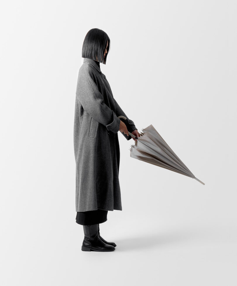 Person in a long gray coat holding a closed umbrella in front of them, standing against a plain white background.