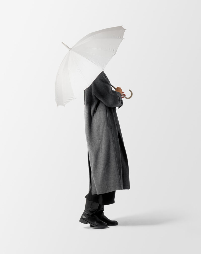 Person in a gray coat and black boots holding a white umbrella, standing against a plain background.