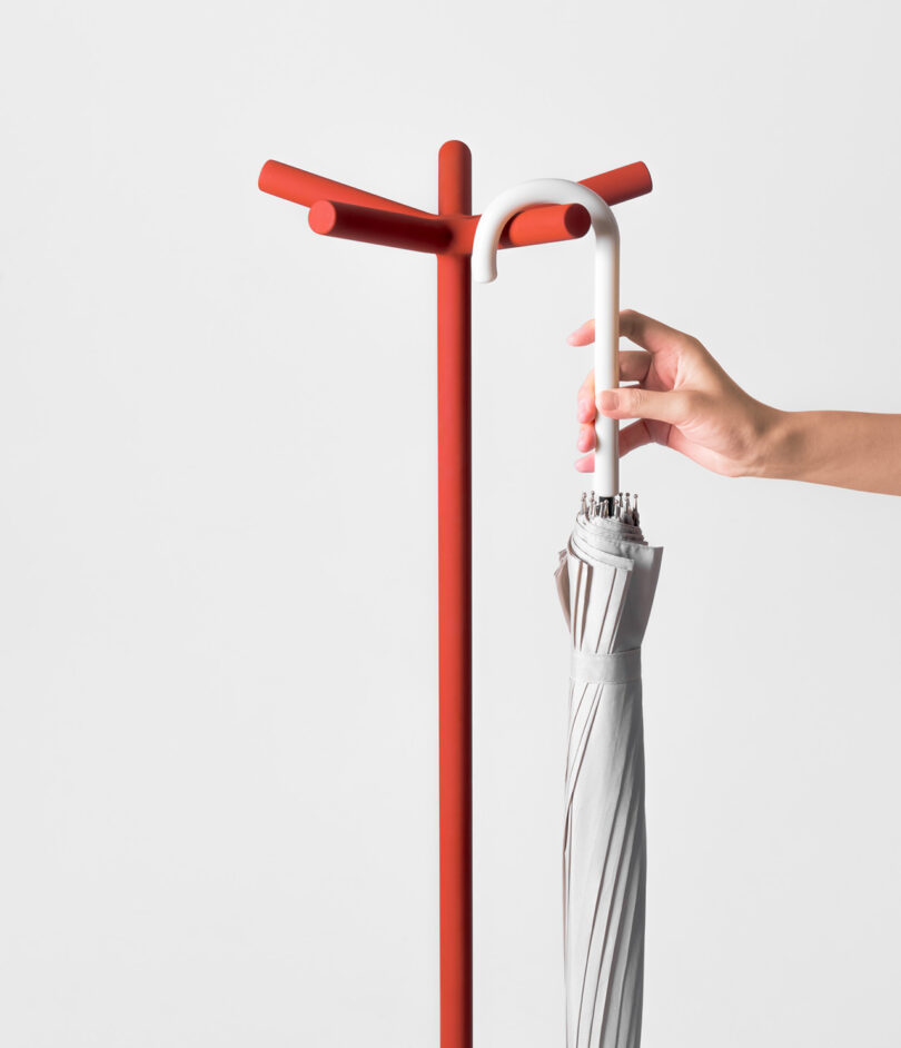 A hand places a closed umbrella on a red, minimalist coat stand with three branches, against a plain white background.