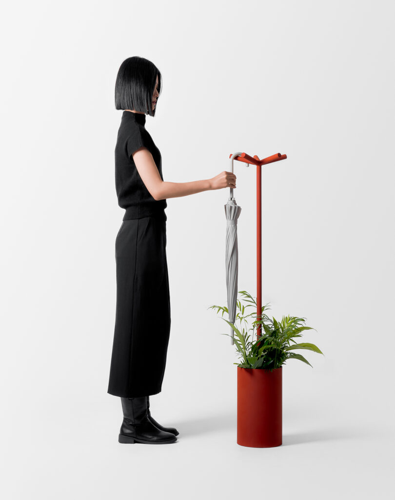 A person in black attire holds a closed umbrella, standing next to a red umbrella stand with green plants.