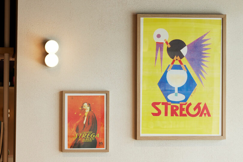 Two framed vintage posters grace the wall; one with a yellow background and an abstract bird design, the other depicting a woman against a red backdrop. These art pieces serve as objects for thought, inviting viewers to engage with their subtle narrative depth.