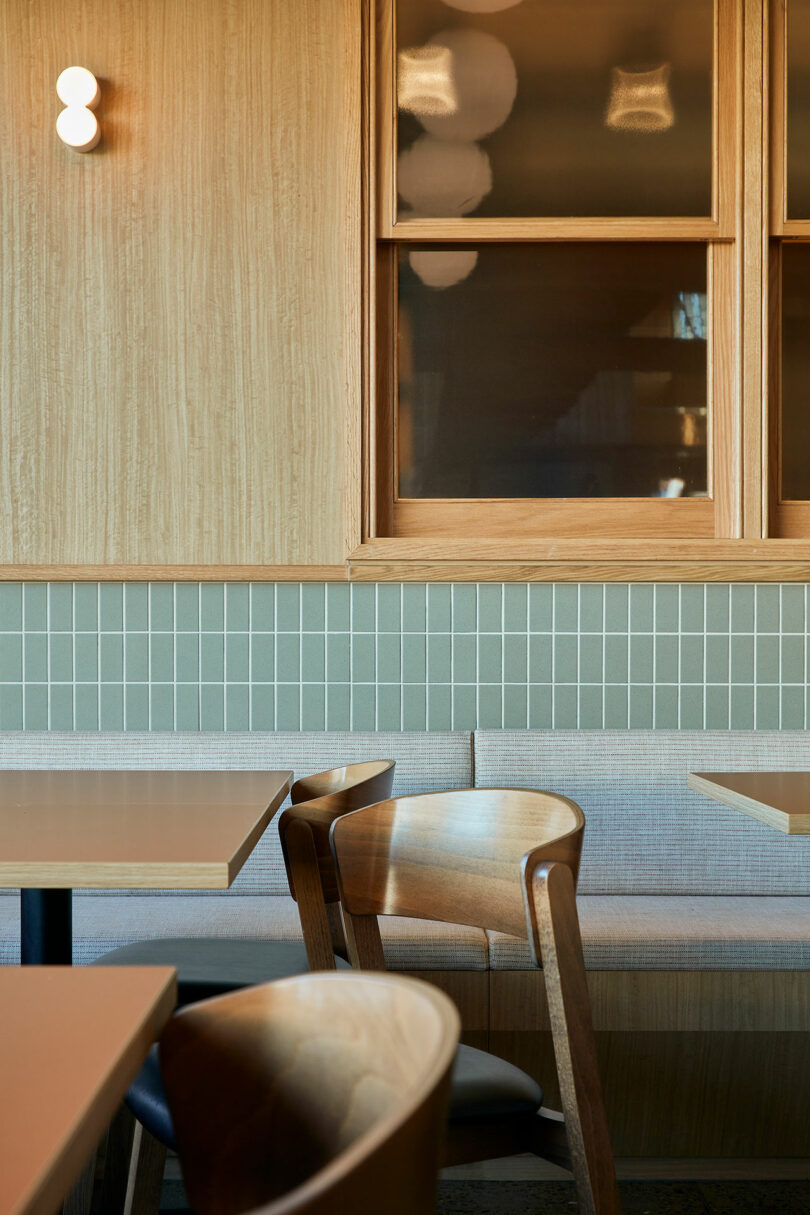 The empty cafe interior, adorned with wooden tables and chairs, features a cushioned bench against a tiled wall. Under the warm light, it serves as an inviting space for patrons to ponder Rider Objects for Thought amidst its serene ambiance.