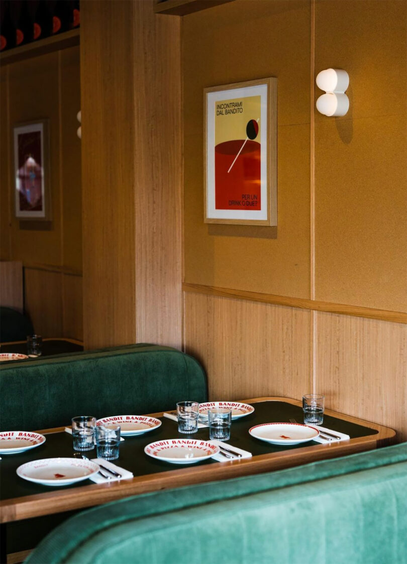 A restaurant booth with green seating, set for four with plates, glasses, and chopsticks. A Rider Objects for Thought poster and a sconce adorn the wood-paneled wall, adding an eclectic touch to the intimate setting.