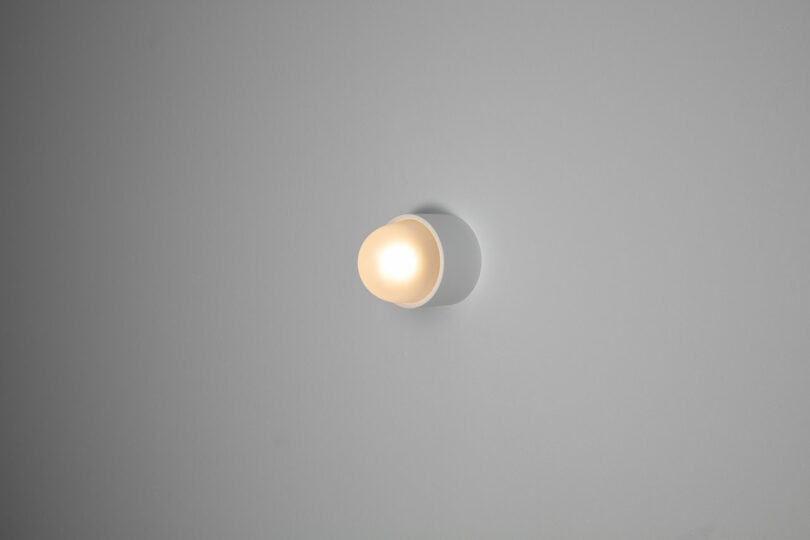 A small, round wall light fixture from the Rider Objects collection emits a soft, warm glow against a plain gray wall, inviting thought and reflection.