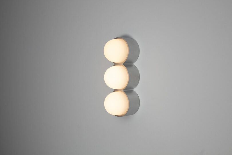 A modern wall light fixture by Rider Objects for Thought features three round, glowing bulbs aligned vertically on a gray wall.