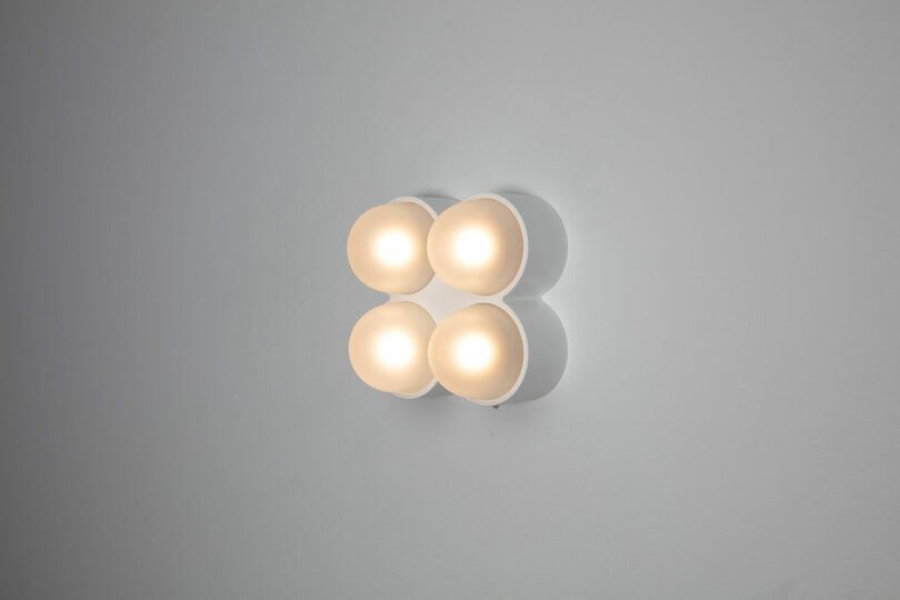 A wall-mounted light fixture with four glowing, rounded bulbs arranged in a square pattern against a gray background serves as an elegant Rider object for thought.