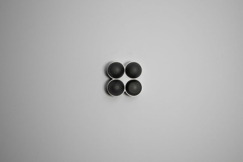 Four small, round black bowls, reminiscent of Rider Objects for Thought, are arranged in a square pattern on a plain gray background.