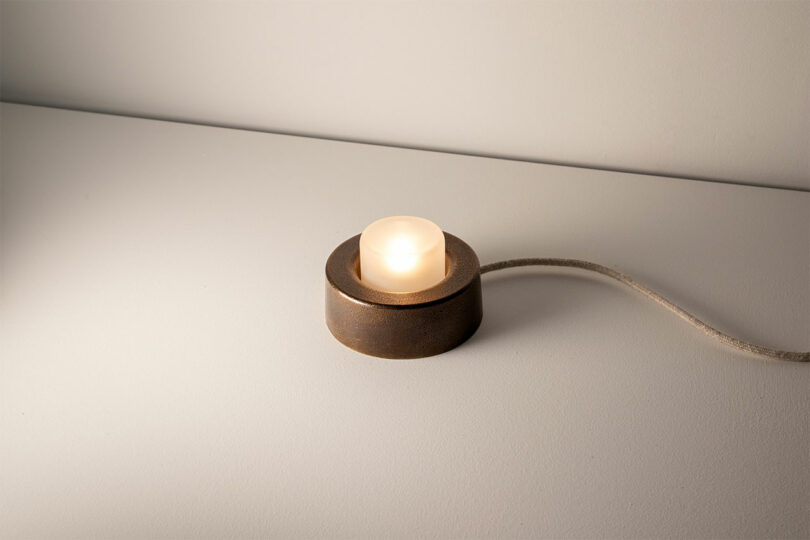 A small, round brown metal light with a white bulb and a cord, reminiscent of Rider Objects for Thought, rests elegantly on a gray surface.