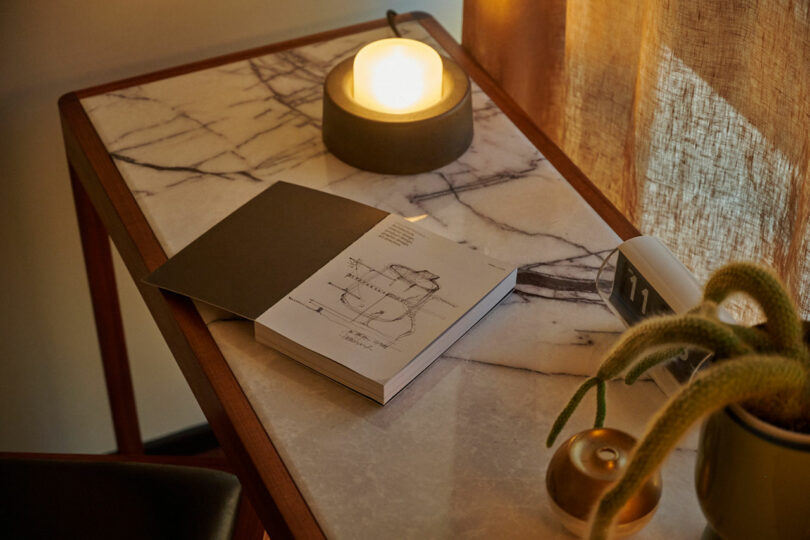 A book with a white cover lies open on a marble-topped table next to a lit round lamp, a small plant, and a calendar displaying "11," as if inviting the rider to pause and ponder these objects for thought.