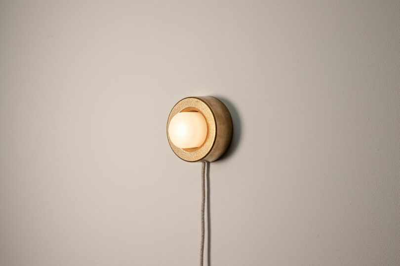 A round wall-mounted light fixture from Rider emits a soft glow against a plain wall, its visible cord adding an element of Objects for Thought to the space.