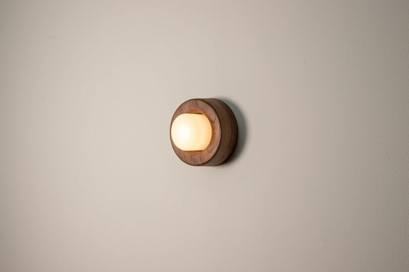 Introducing the Rider Objects for Thought collection: a round, wooden wall-mounted lamp that beautifully frames a glowing white bulb against a plain light wall.